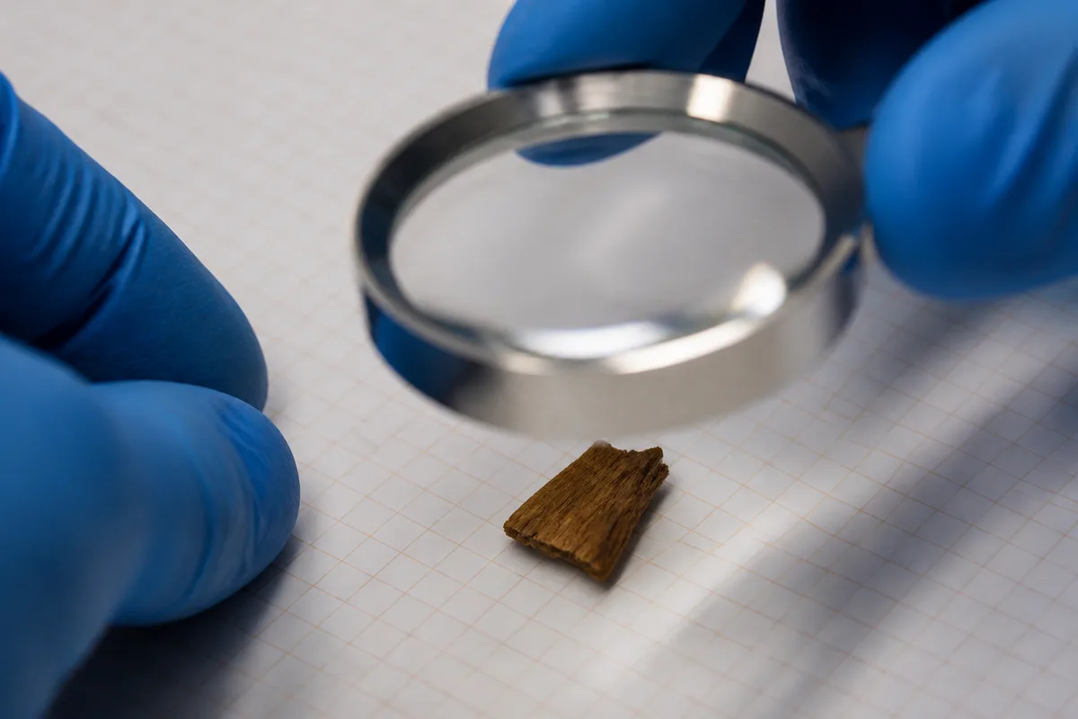 Mains gantées approchant une loupe d&rsquo;un fragment de résine brune posé sur un carnet quadrillé